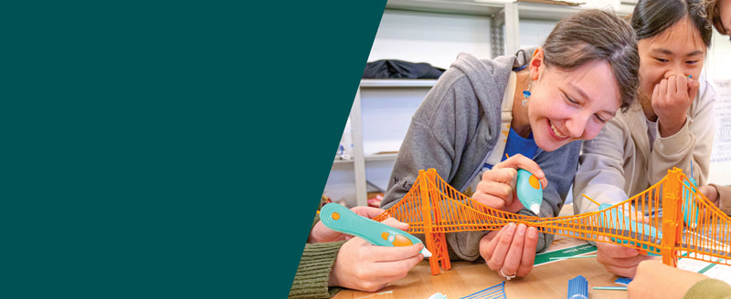Three people smiling and building a colorful orange suspension bridge model using 3D printing pens on a table in a bright indoor space.